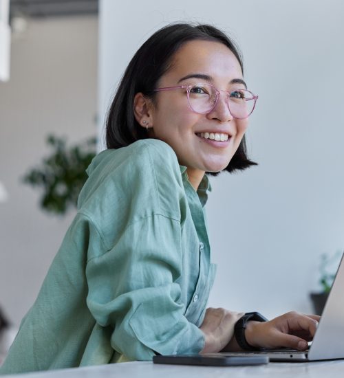 Pretty cheerful Asian woman in eyeglasses and casual clothes browses laptop computer connected to 4g internet updates software uses modern technologies poses in cafeteria looks gladfully away
